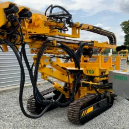 Żółta, gąsienicowa wiertnica Beretta T59 z licznymi przewodami hydraulicznymi, ustawiona na żwirowym podłożu, z widocznym tłem zabudowań i drzew