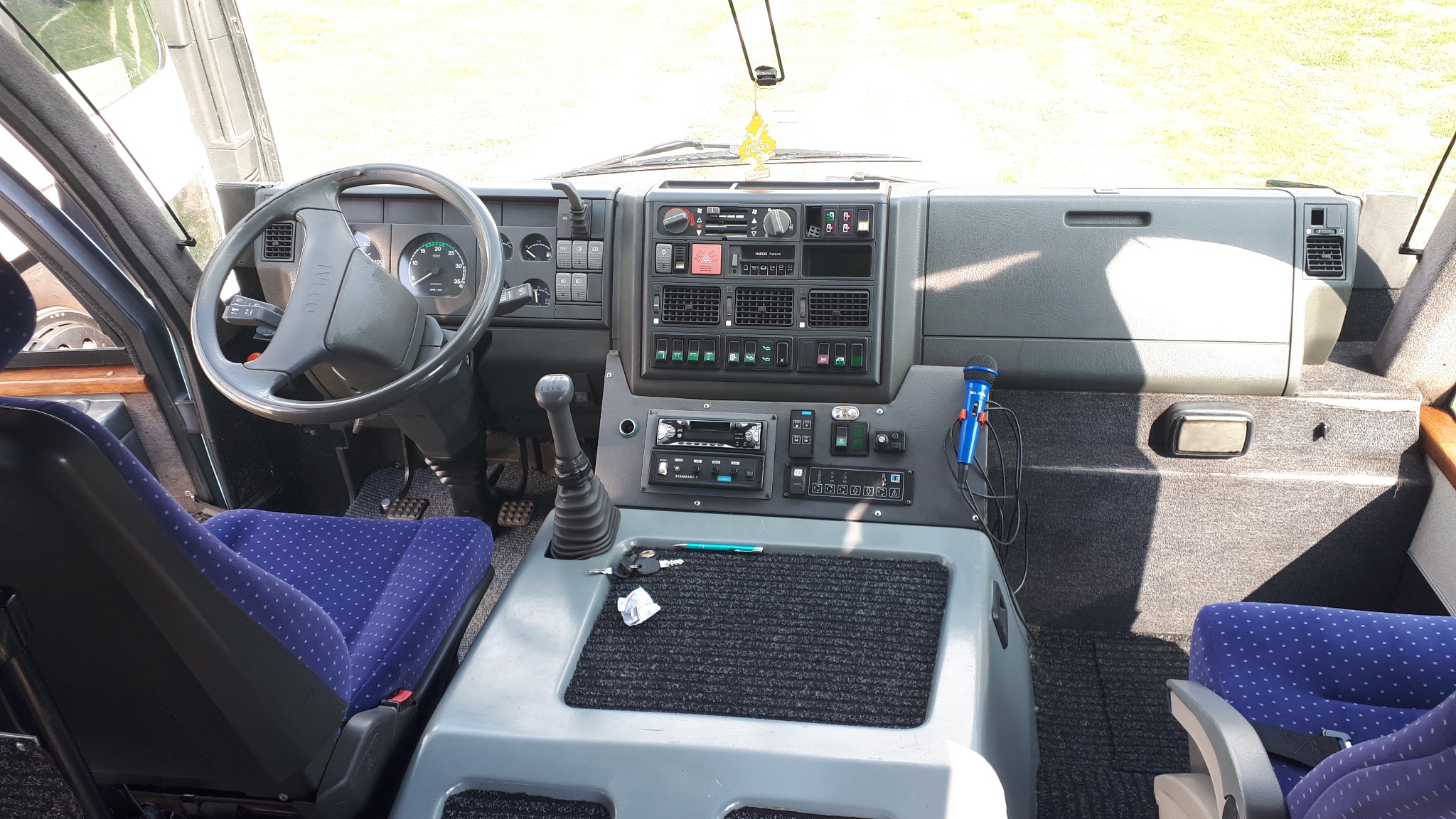 Wnętrze autobusu z widoczną deską rozdzielczą, kierownicą, mikrofonem i niebieskimi fotelami z białymi kropkami.