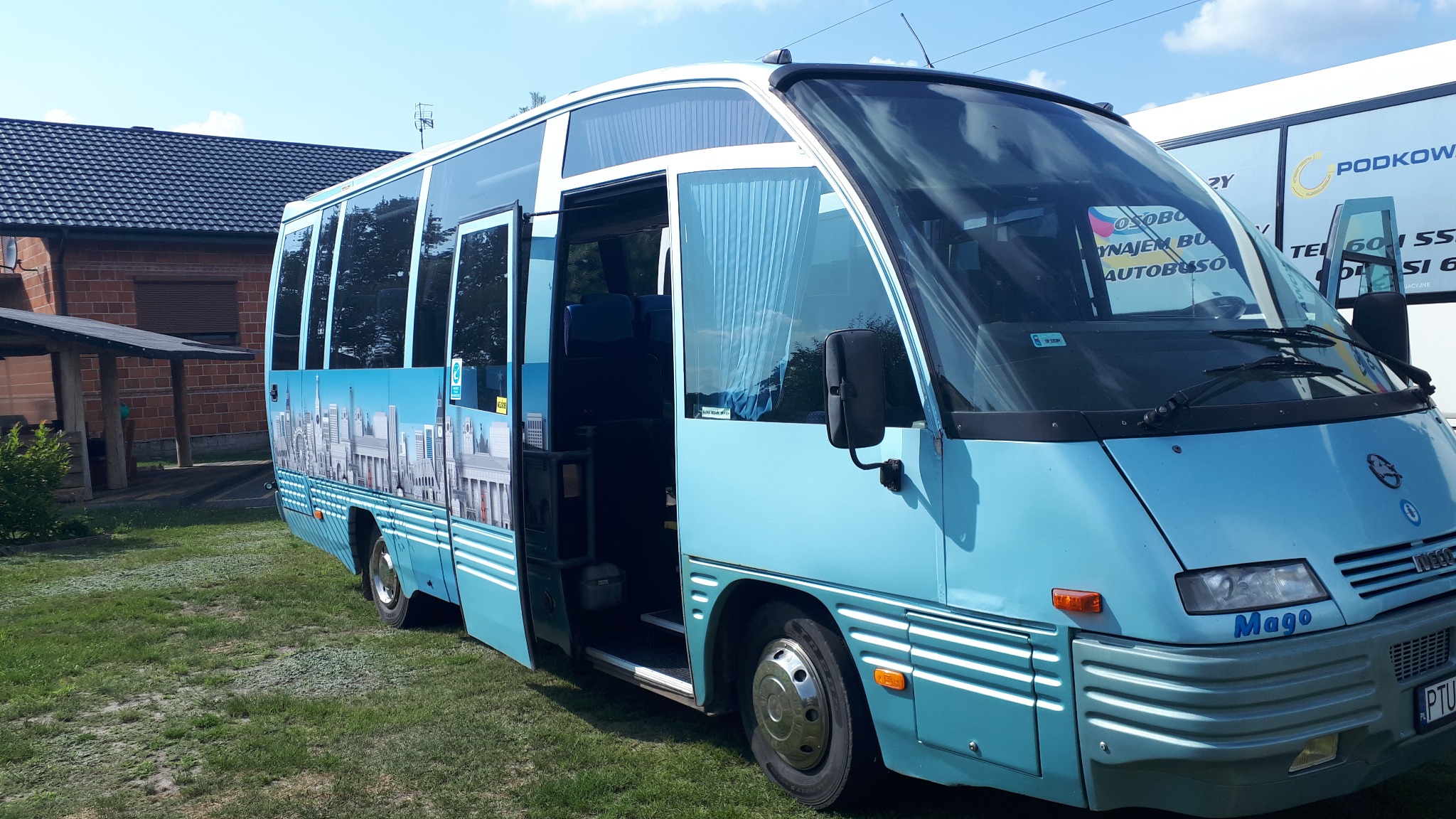 Niebieski bus Iveco Mago z otwartymi drzwiami, dekorowany grafiką panoramy miasta, zaparkowany na trawie.