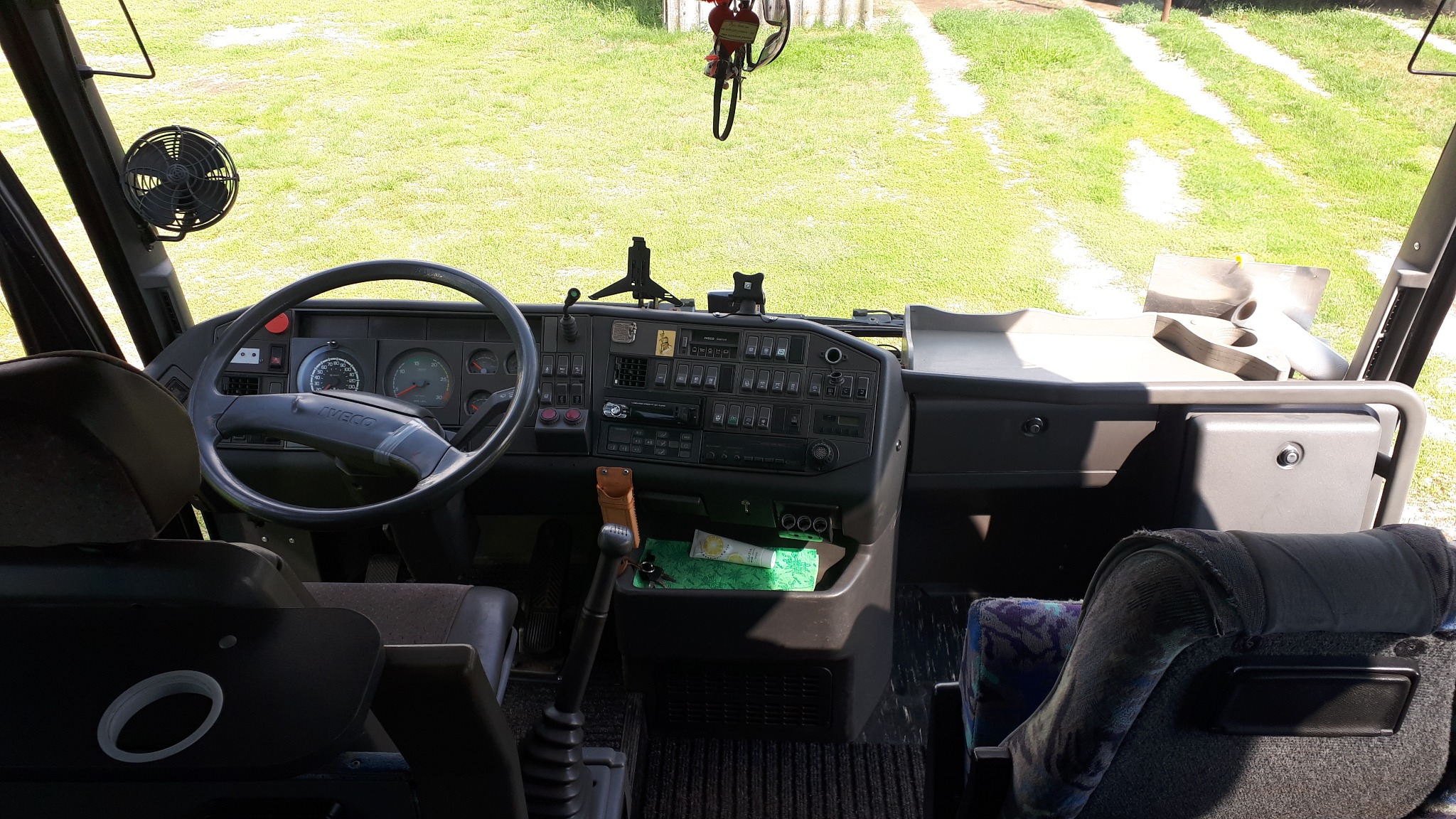 Wnętrze autobusu marki Iveco z widokiem na deskę rozdzielczą, kierownicę i wentylator, z zewnątrz widoczna trawiasta droga.
