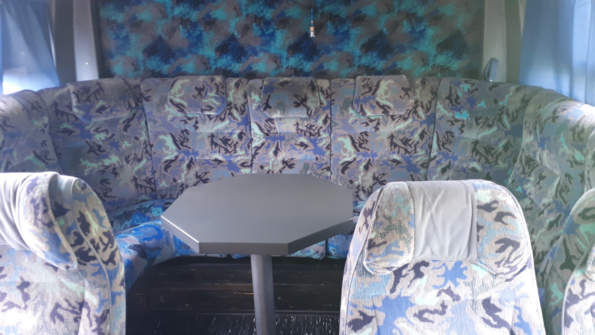 Wnętrze autobusu z tapicerką w odcieniach niebieskiego, szarego i zielonego, z widocznym stolikiem o ośmiokątnym blacie.