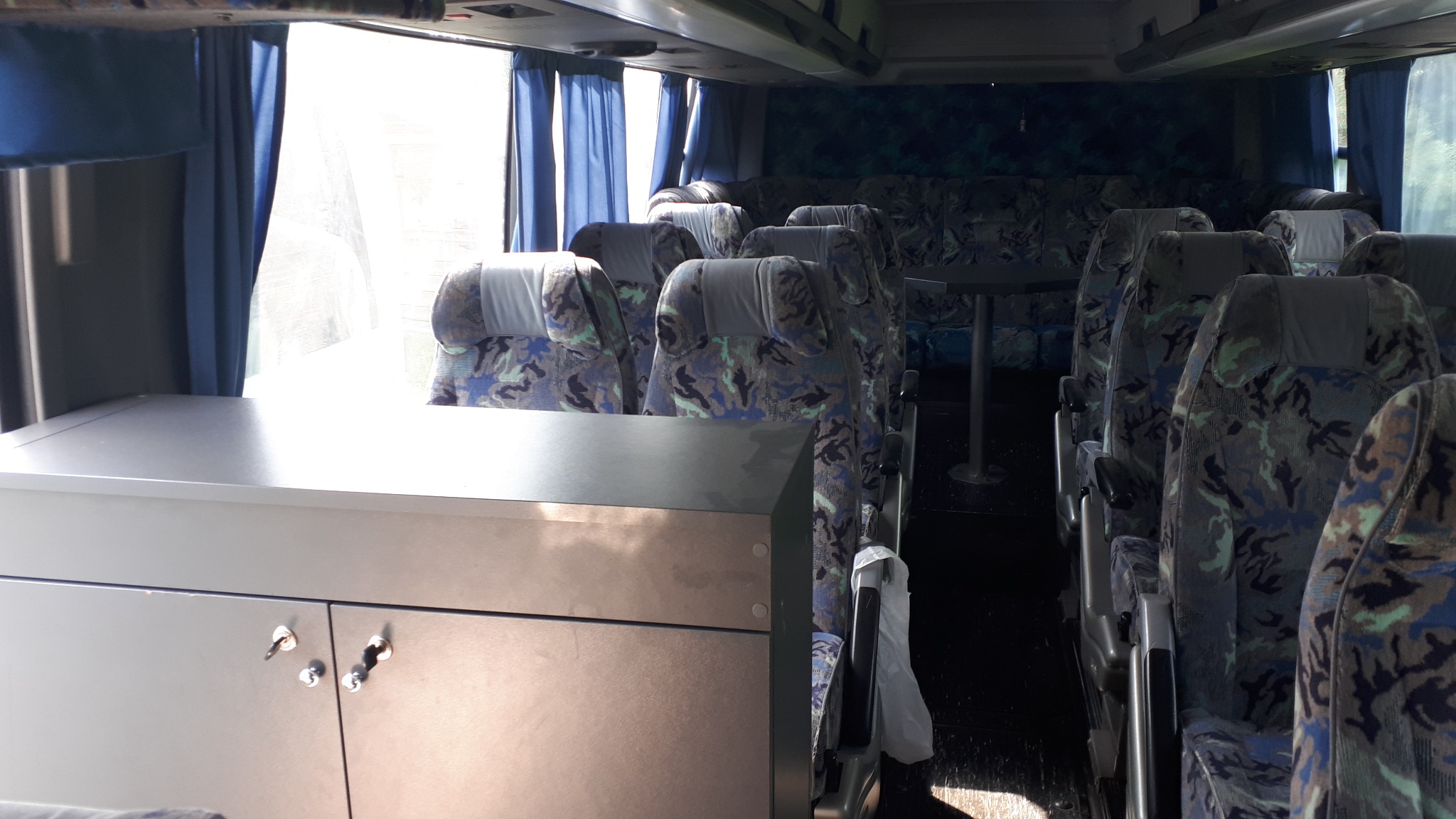 Wnętrze autobusu turystycznego z rzędami foteli w kamuflażu i zasłonami przy oknach, widoczna szafka z kluczami.