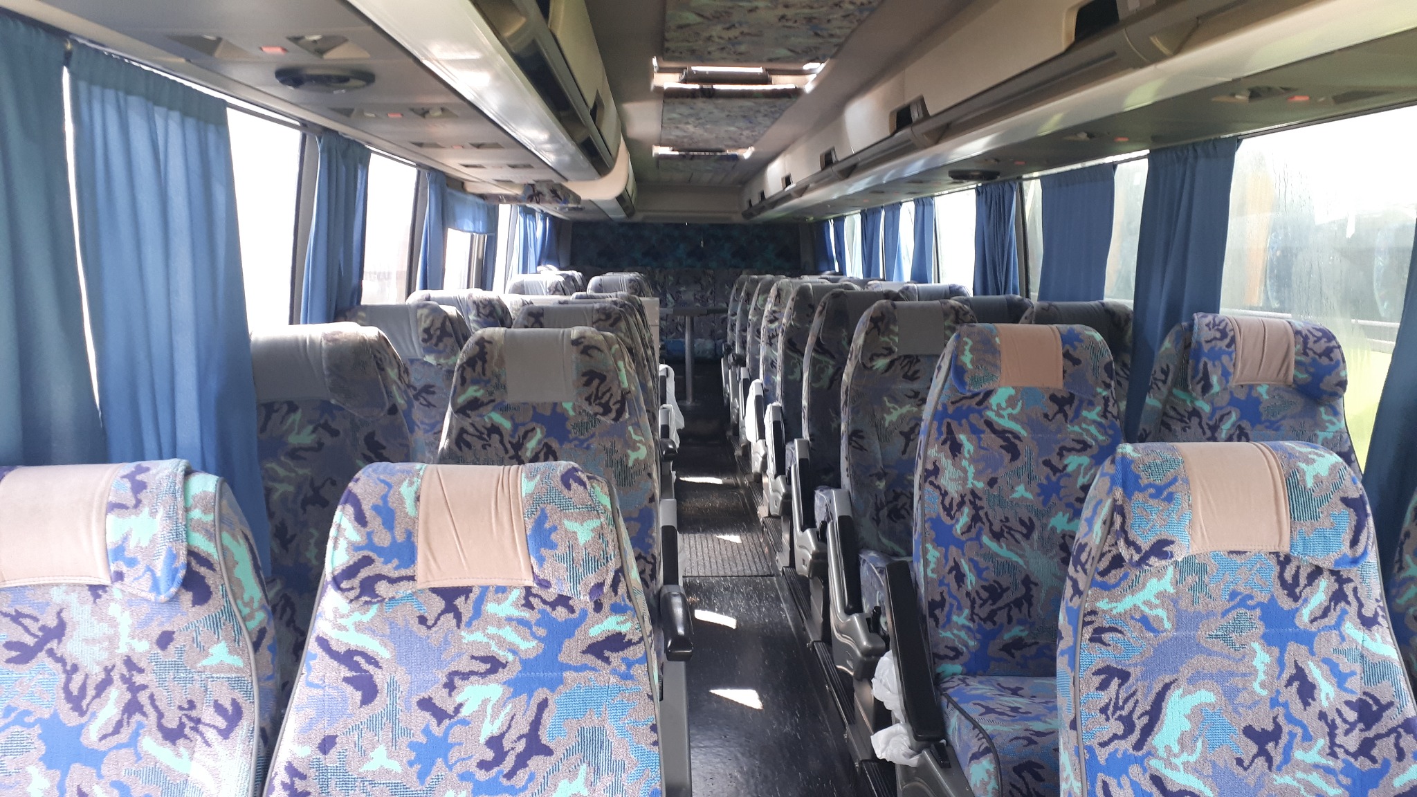Wnętrze autobusu z rzędami siedzeń w niebiesko-szare wzory kamuflażu, jasne zasłony przy oknach, widok z przodu pojazdu.
