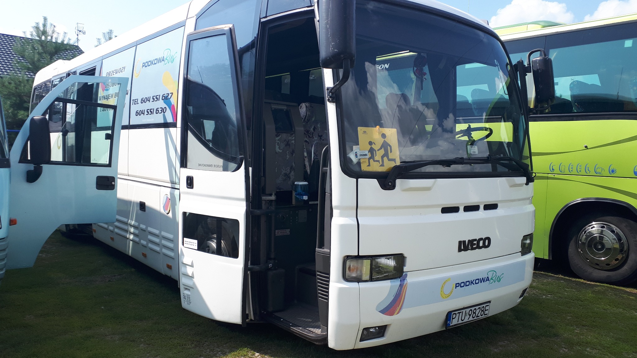 Biały autobus Iveco z otwartymi drzwiami wejściowymi, widoczna naklejka 'Uwaga dzieci' na przedniej szybie, logo 'Podkowa Bus' na karoserii, zaparkowany na trawie.