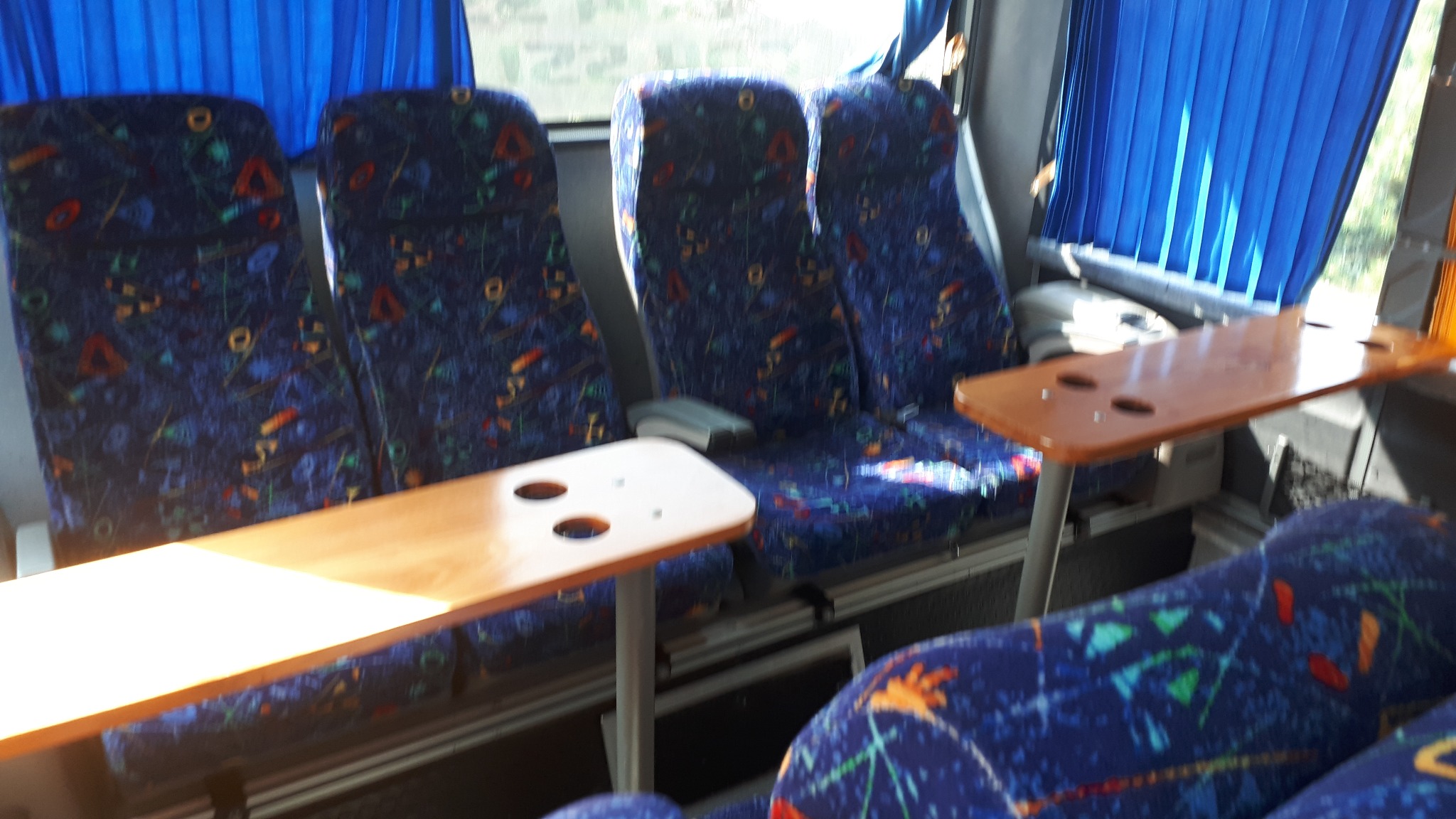 Wnętrze autobusu z niebieskimi fotelami w geometryczne wzory i drewnianymi stolikami przy oknach zasłoniętych niebieskimi zasłonami.