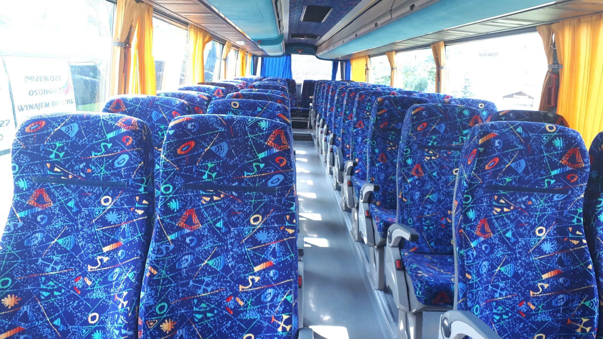 Wnętrze autobusu z rzędami niebieskich foteli z geometrycznym wzorem, widok z przodu pojazdu, żółte zasłony w oknach.