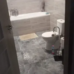 Jasna łazienka z wanną, toaletą i szarą marmurową podłogą z wzorem; widoczna butelka płynu do czyszczenia pod toaletą.