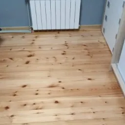 Jasna podłoga z desek sosnowych z widocznymi sękami, grzejnik żeberkowy pod oknem z roślinami doniczkowymi, fragment białej komody.