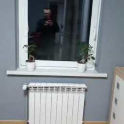 Szare wnętrze pokoju z białym oknem, grzejnikiem i drewnianą podłogą. W oknie odbija się osoba robiąca zdjęcie telefonem.