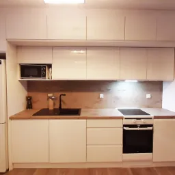 Jasna kuchnia z zabudową pod sufit, drewniany blat, czarny zlew i płyta grzewcza, widoczna lodówka i mikrofalówka.