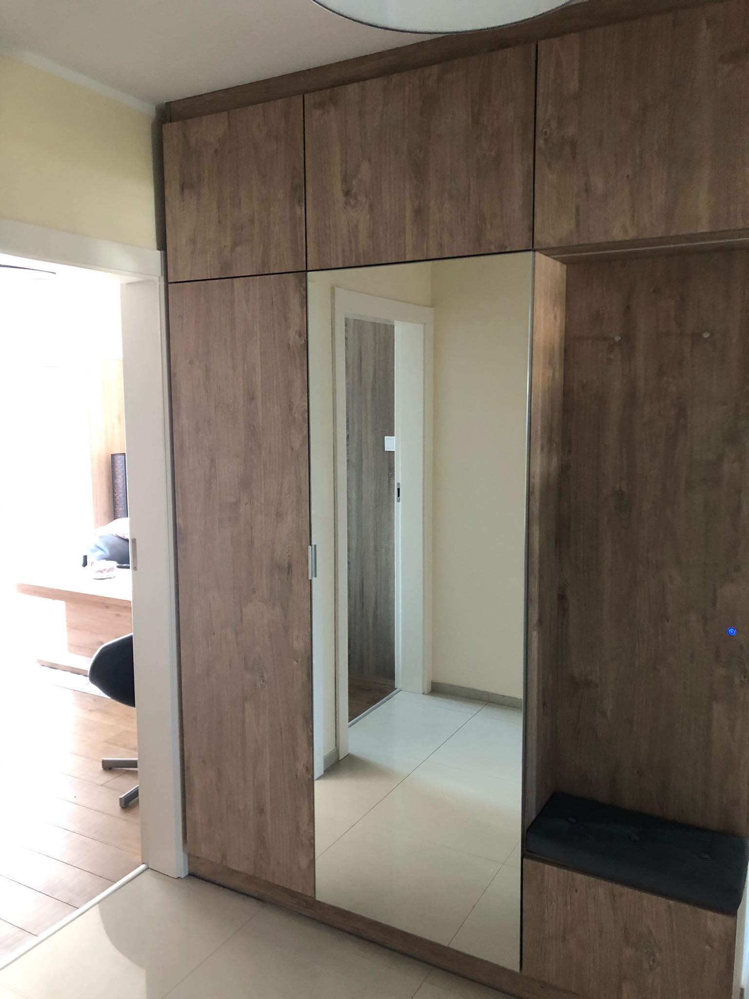 Wbudowana szafa z lustrem i siedziskiem w przedpokoju, drewnopodobne wykończenie, widoczne odbicie wnętrza mieszkania.