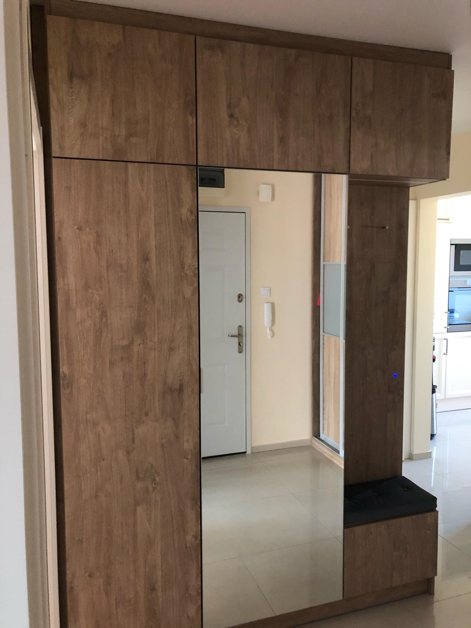 Szafa na wymiar z lustrem i siedziskiem w przedpokoju, fornir w odcieniach brązu z widocznym rysunkiem drewna, na ścianie białe drzwi wejściowe.