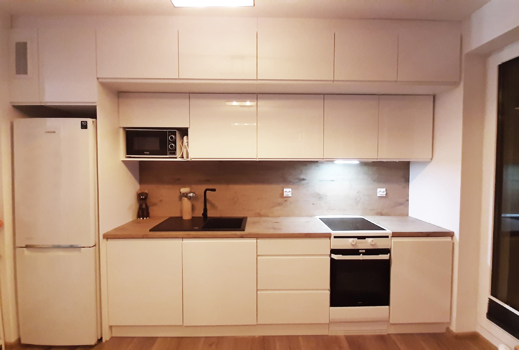 Jasna kuchnia z zabudową pod sufit, drewniany blat, czarny zlew i płyta grzewcza, widoczna lodówka i mikrofalówka.