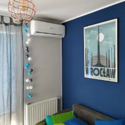 Biały klimatyzator Haier zamontowany w szarym pokoju z niebieską ścianą, ozdobiony lampą w stylu industrialnym, girlandą świetlną z kulkami oraz plakatem z motywem Wrocławia.