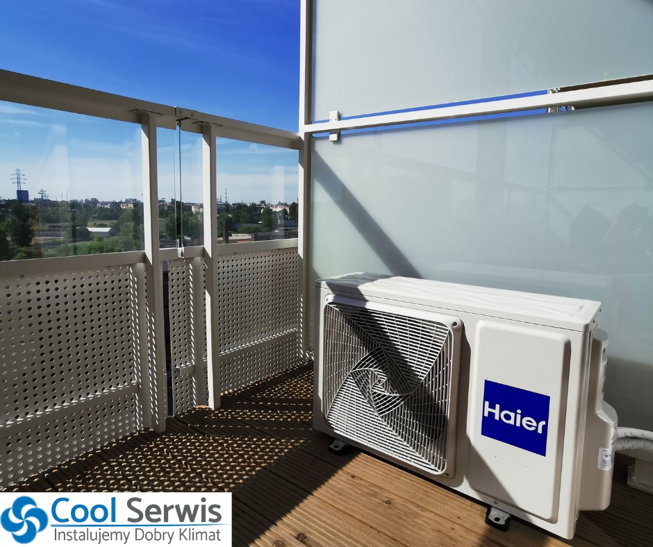 Jednostka zewnętrzna klimatyzacji Haier na balkonie z drewnianą podłogą, widok na miasto w tle.
