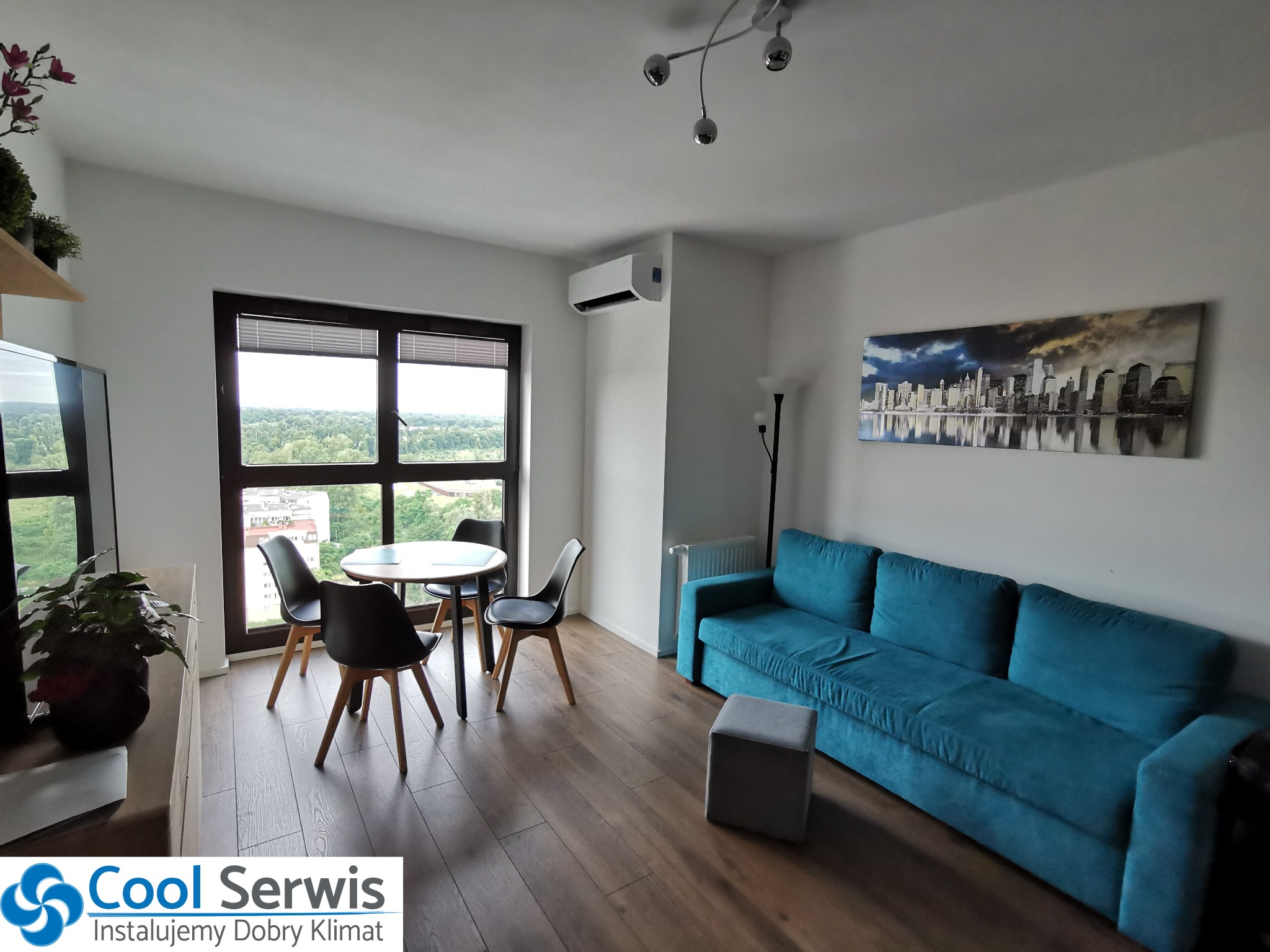 Wnętrze salonu z zamontowanym klimatyzatorem ściennym nad oknem, widok na stół z krzesłami, turkusową sofę i obraz z panoramą miasta.