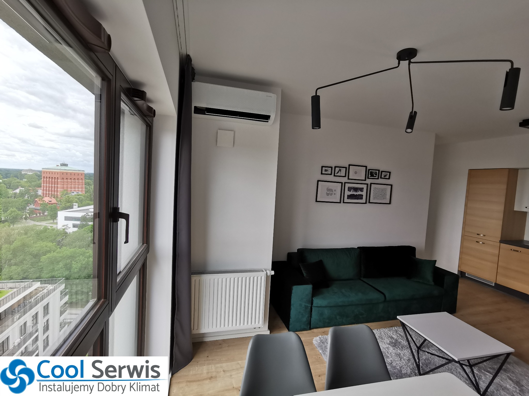 Nowoczesny salon z zamontowanym klimatyzatorem ściennym Samsung, widok z okna na zielone otoczenie miejskie, szary zasłona i designerska lampa sufitowa.