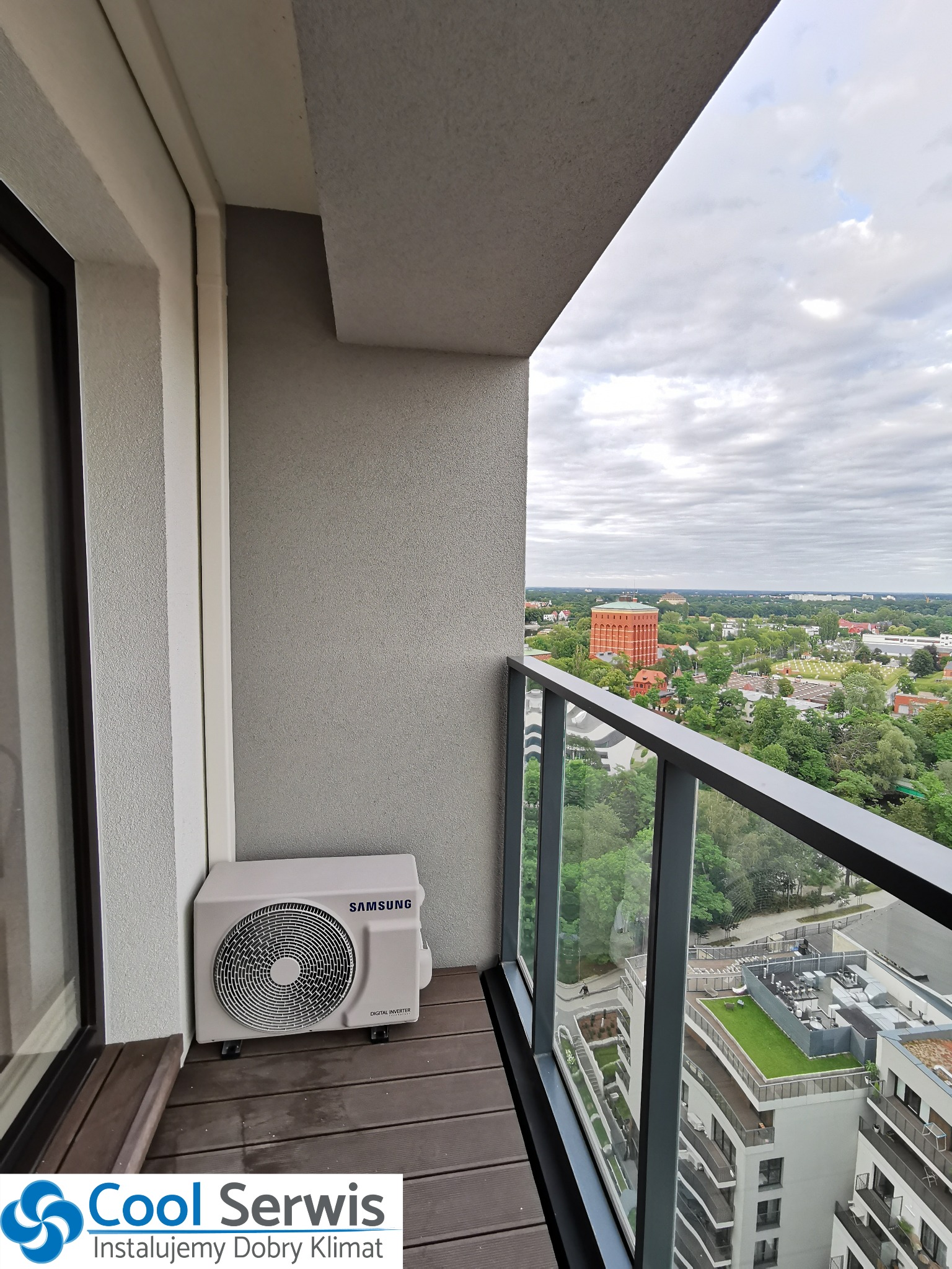 Zewnętrzna jednostka klimatyzacji Samsung Digital Inverter na drewnianym balkonie z widokiem na miasto i wieżę ciśnień w tle.