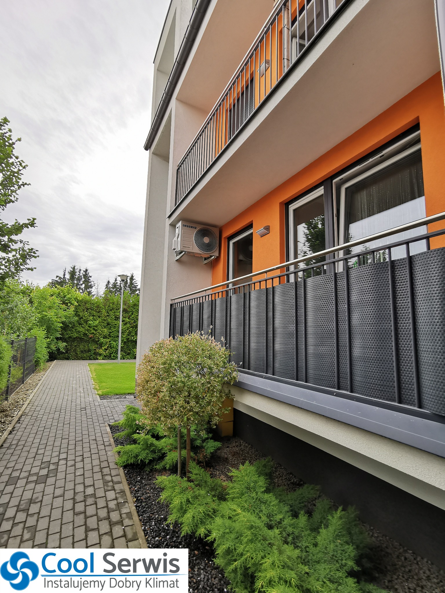 Zewnętrzna jednostka klimatyzacji Samsung zamontowana na elewacji budynku wielorodzinnego z pomarańczowym balkonem, widoczna z poziomu chodnika z kostki brukowej.