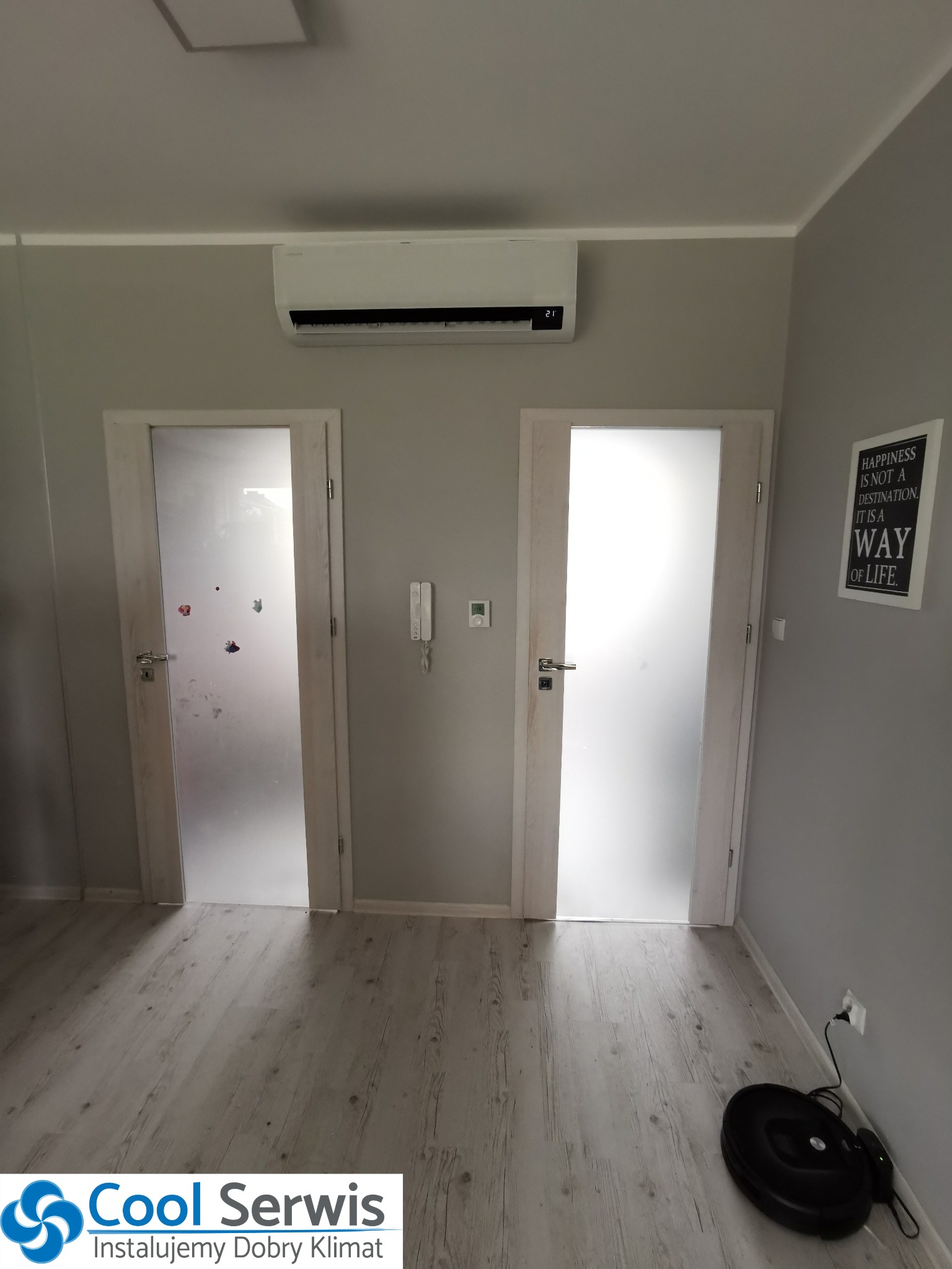 Biały klimatyzator ścienny zamontowany nad dwoma drzwiami z matowego szkła w szarym pomieszczeniu mieszkalnym, z robotem sprzątającym przy ścianie i dekoracyjnym plakatem.