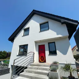 Nowoczesny dom z białą elewacją, czarnym dachem i czerwoną stolarką drzwiową, widok z dołu na wejście z roślinami ozdobnymi.