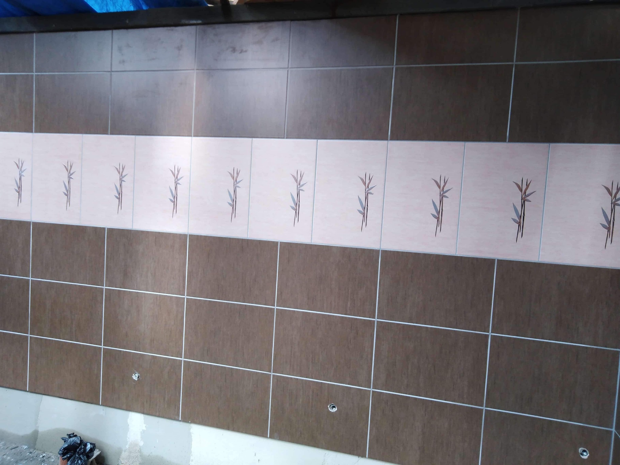 Ułożone brązowe i beżowe płytki ścienne z motywem roślinnym, widoczne otwory na armaturę, fragment worka na gruz przy podłodze.