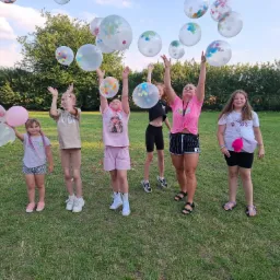 Grupa dzieci i dorosłych na trawie, radośnie podrzucających do góry przezroczyste balony wypełnione kolorowym konfetti.