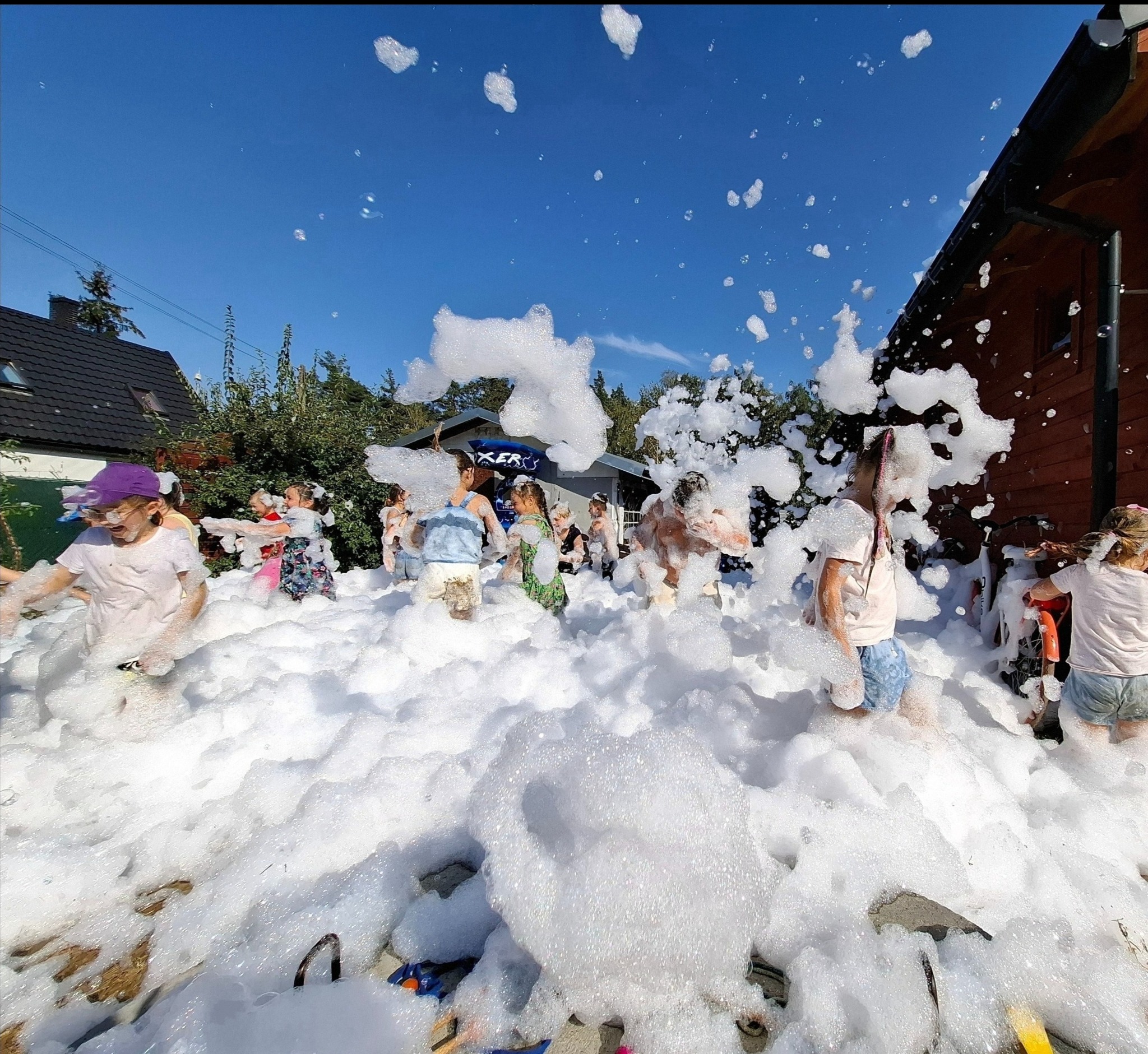 Grupa dzieci bawi się w pianie podczas imprezy plenerowej, widoczne budynki w tle i czyste niebo.