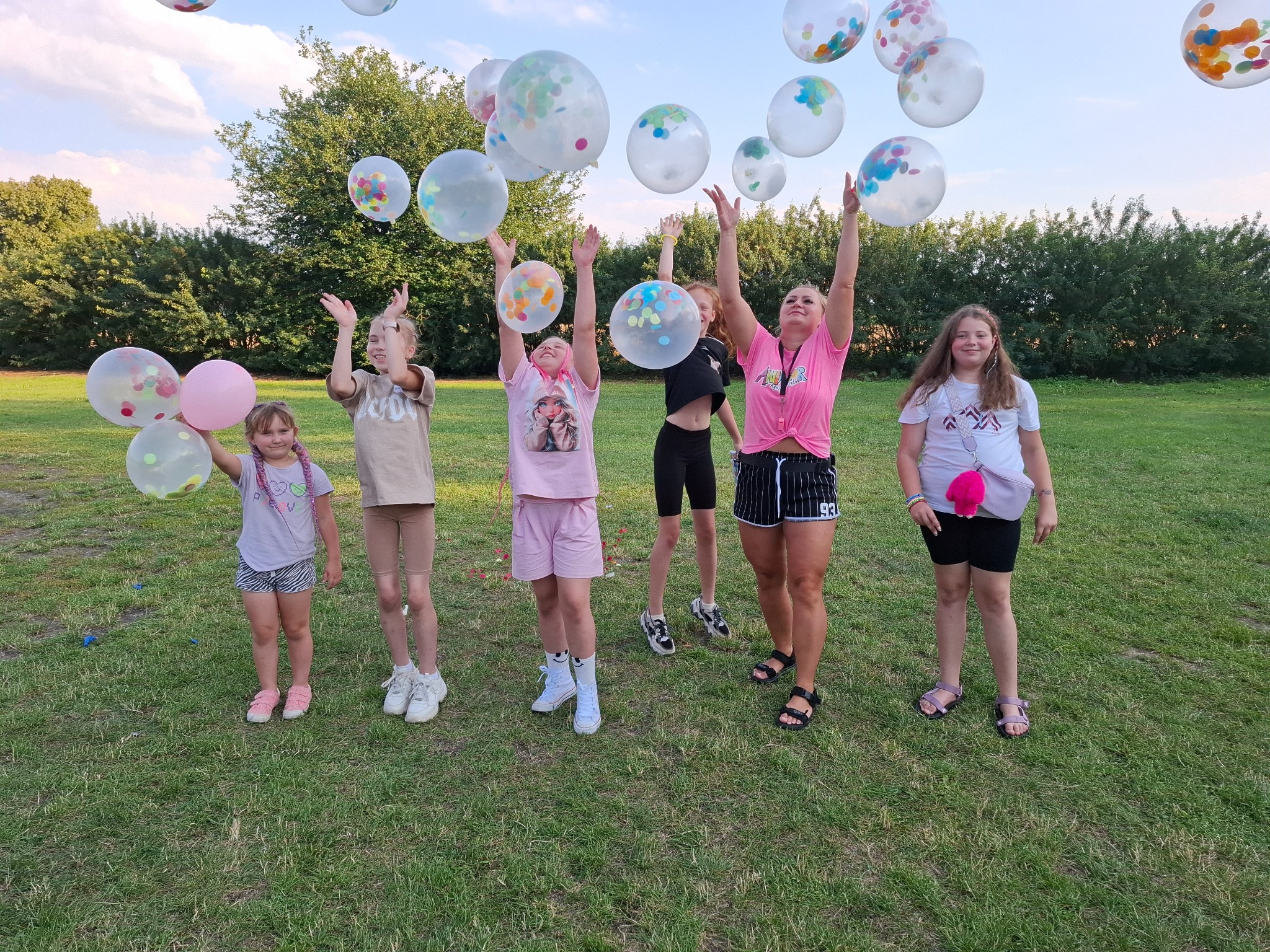 Grupa dzieci i dorosłych na trawie, radośnie podrzucających do góry przezroczyste balony wypełnione kolorowym konfetti.