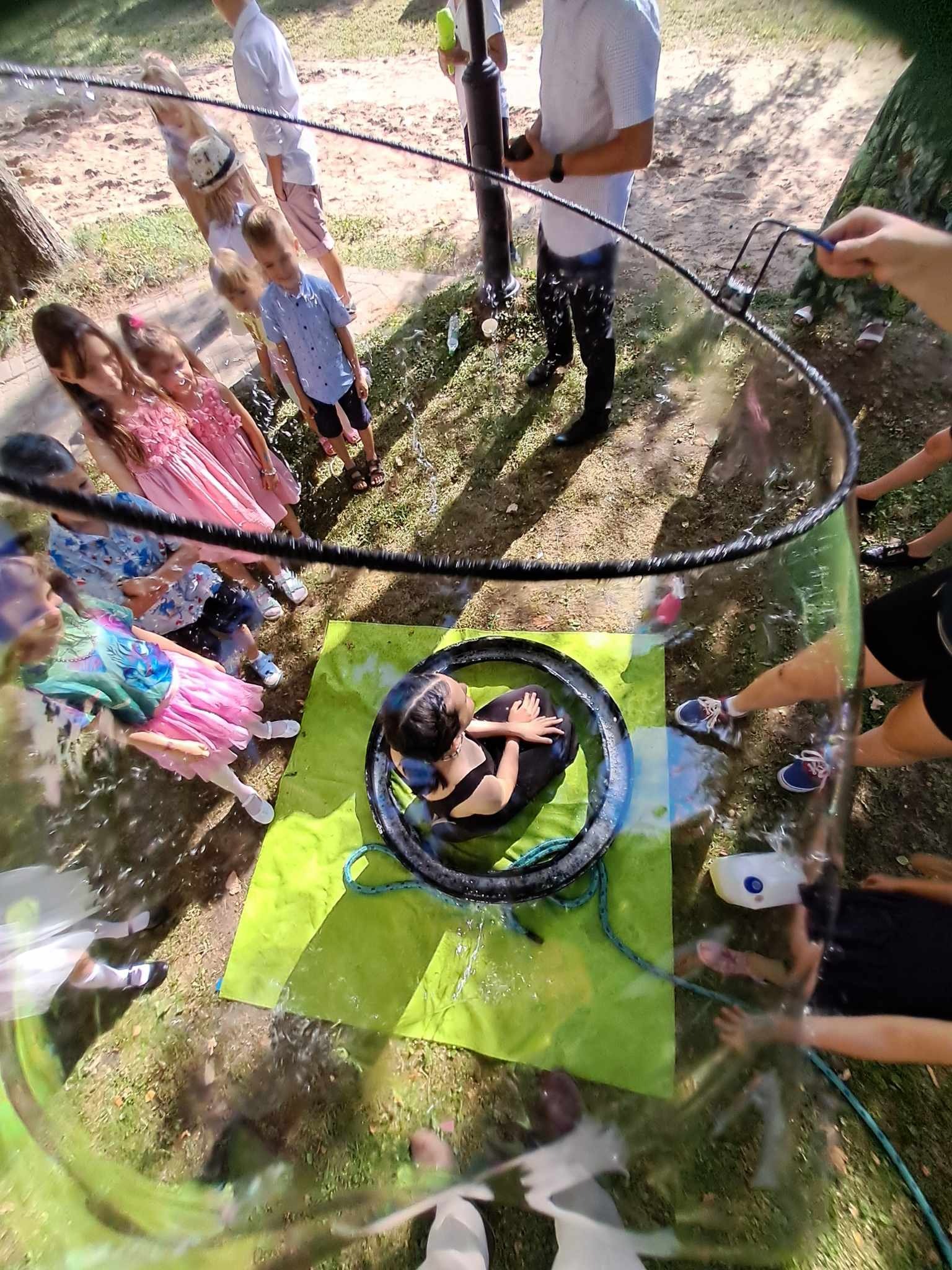 Dziewczynka w czarnej sukience siedzi wewnątrz dużej bańki mydlanej, widok z góry, dzieci obserwują, zielona mata pod spodem.