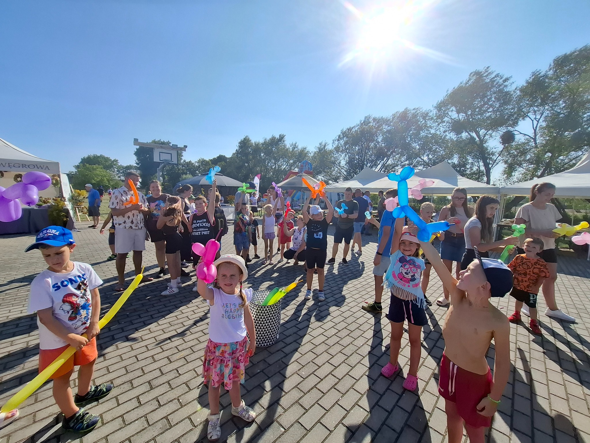 Grupa dzieci trzymających balony w różnych kształtach podczas imprezy plenerowej, widoczne namioty i dekoracje.