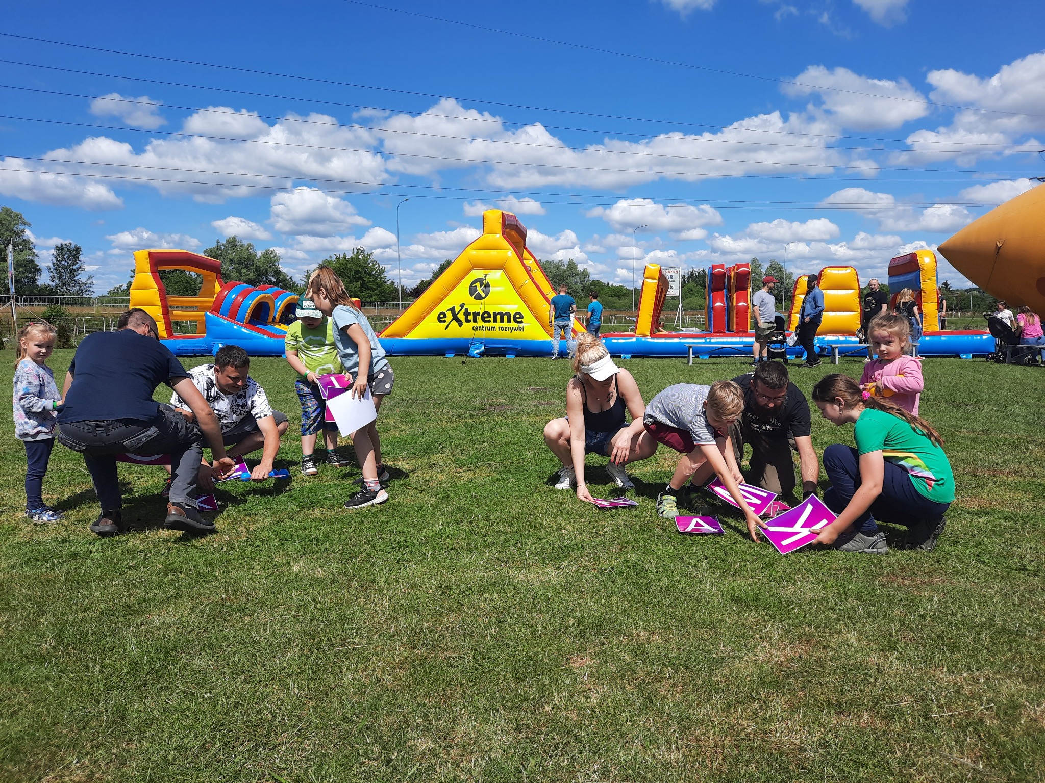 Grupa dzieci i dorosłych układa litery z dużych kartonowych elementów na trawie, w tle dmuchany plac zabaw z logo 'extreme centrum rozrywki', błękitne niebo z chmurami.