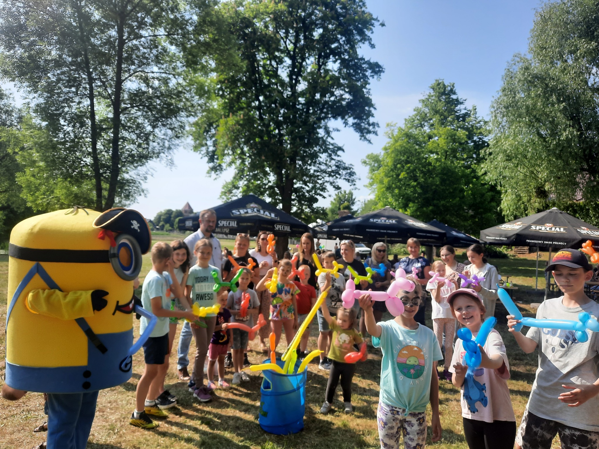 Animator w kostiumie Minionka Pirata i grupa dzieci trzymających balony modelowane w kształcie zwierząt na tle zieleni parkowej podczas imprezy plenerowej.