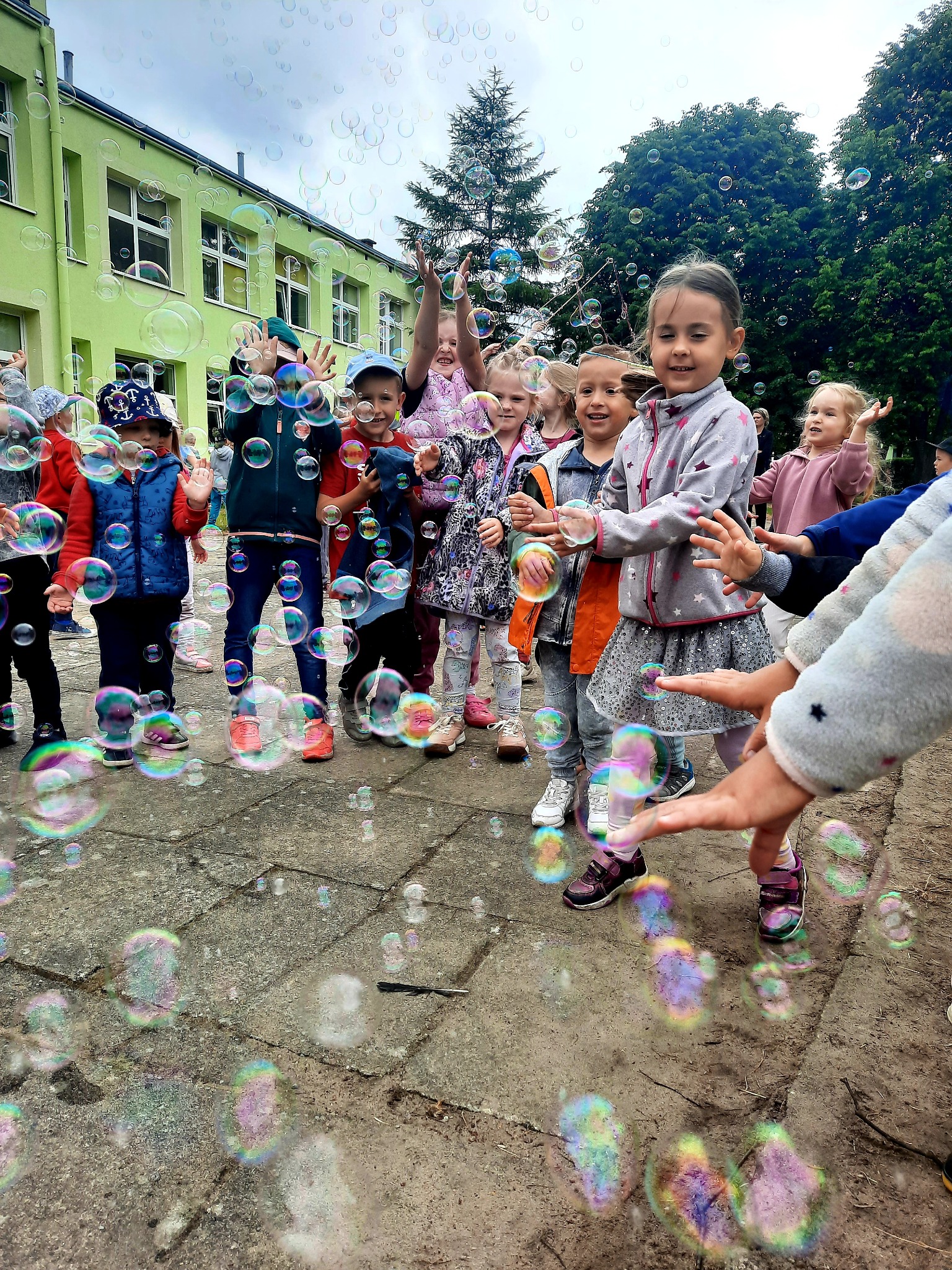 Grupa dzieci bawi się na zewnątrz, łapiąc bańki mydlane na tle zielonego budynku szkolnego i drzew.