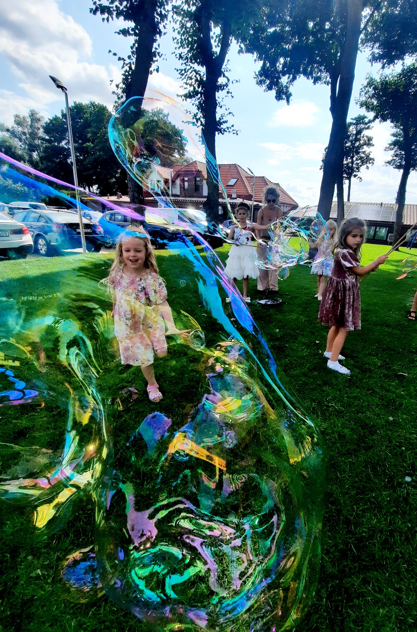 Dziewczynka w kwiecistej sukience biegnie przez trawnik, otoczona ogromnymi, opalizującymi bańkami mydlanymi, tworzonymi przez osobę dorosłą i inne dzieci w tle.