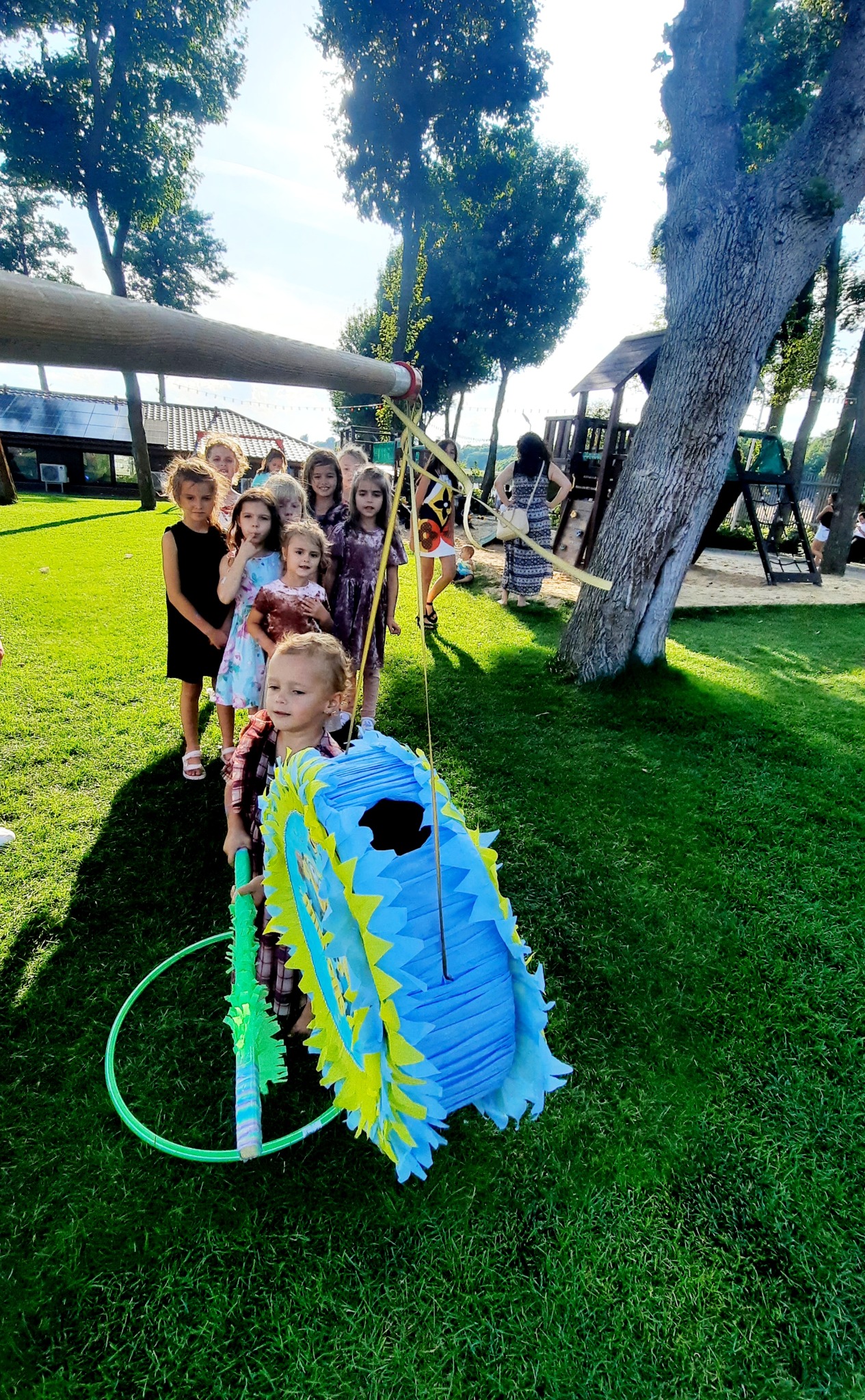 Grupa dzieci ustawiona w kolejce na trawie, czekająca na swoją kolej do rozbicia niebiesko-żółtej piniaty w kształcie gwiazdy, trzymanej przez małego chłopca z zielonym kijem i obręczą.