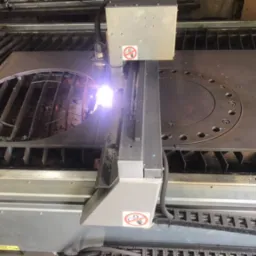 Proces cięcia plazmowego grubej, stalowej płyty z wyciętym okręgiem i otworami na śruby, iskry i dym unoszą się znad obszaru cięcia.