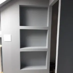 Szara ściana z trzema wnękami, świeżo po malowaniu, widoczny włącznik światła i fragment podłogi zabezpieczonej folią malarską.