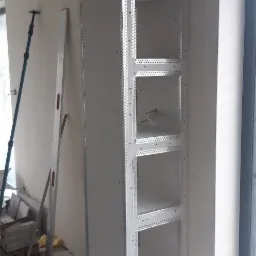 Wnęka ścienna z płyt kartonowo-gipsowych z metalowymi narożnikami, widoczne kable w jednej z półek, poziomica i podpora teleskopowa w tle.