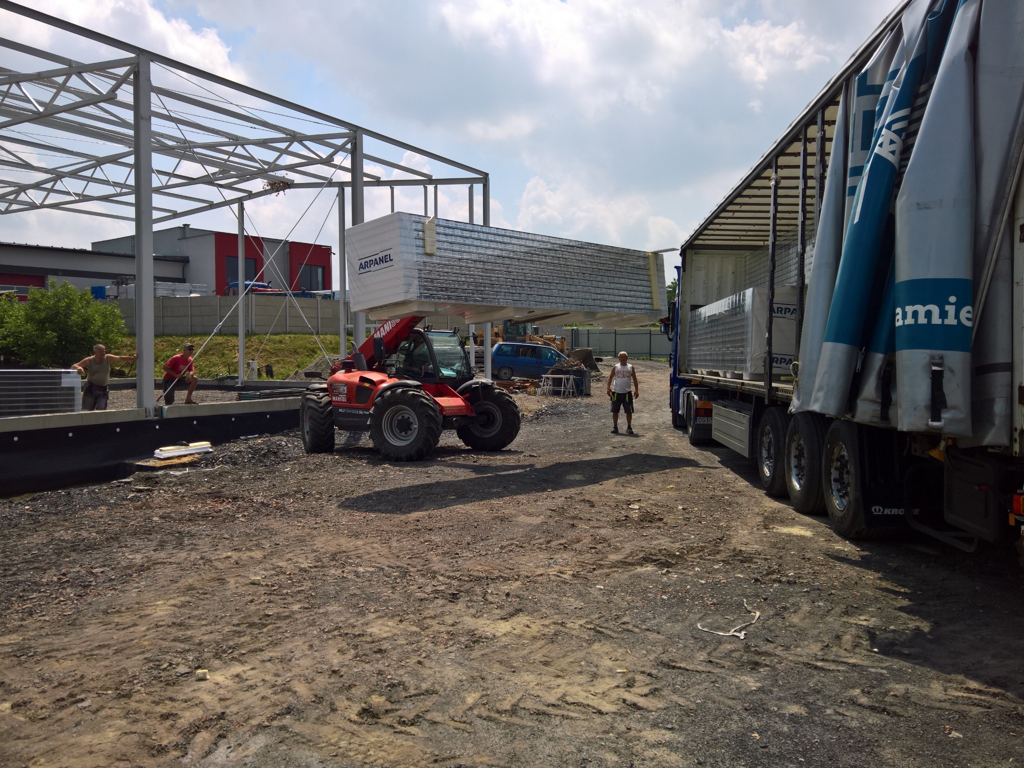 Czerwony wózek widłowy Manitou transportuje pakiet płyt warstwowych Arpanel z naczepy ciężarówki na placu budowy, gdzie wznoszona jest stalowa konstrukcja hali.