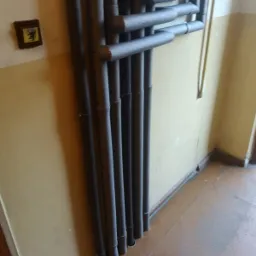 Pionowe rury grzewcze z szarą izolacją termiczną, biegnące wzdłuż żółtej ściany w korytarzu, widoczne także elementy podłogi i fragment framugi drzwi.