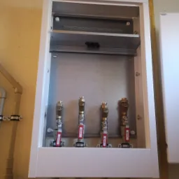 Wnętrze szafki z czterema zaworami kulowymi Calido z czerwonymi rączkami, zamontowanymi na rurach, widoczne elementy instalacji hydraulicznej.