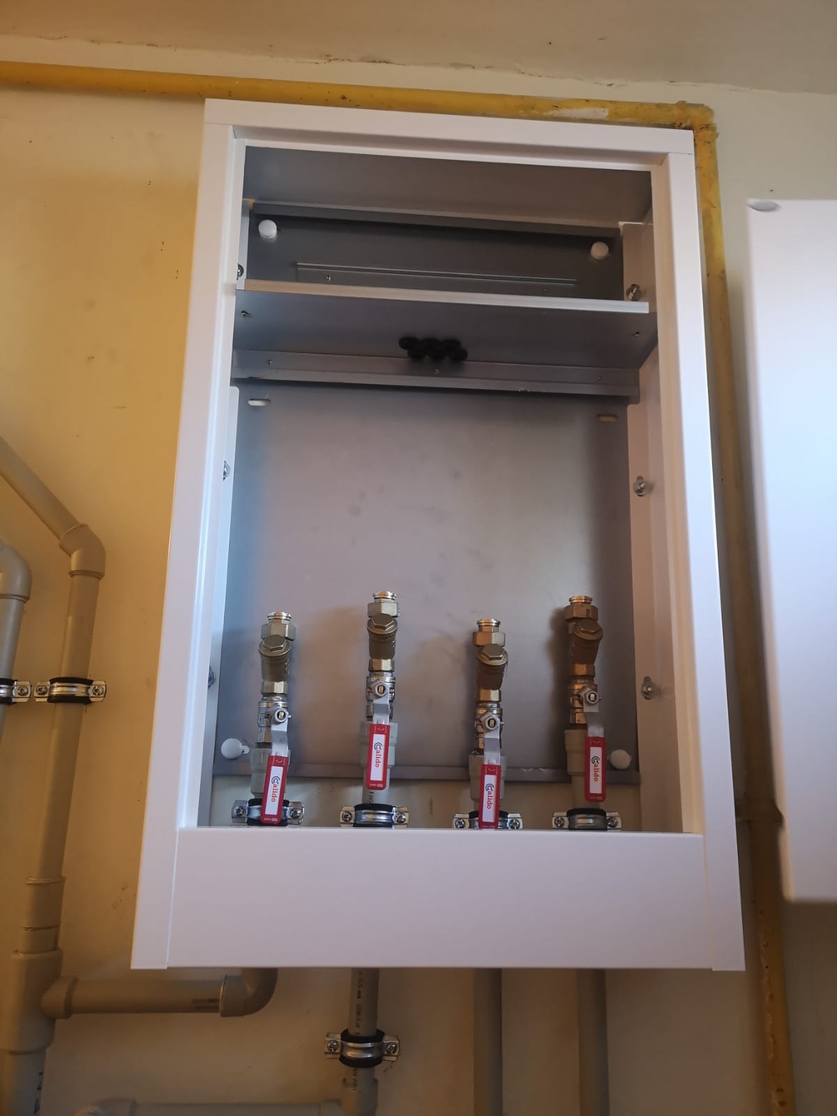 Wnętrze szafki z czterema zaworami kulowymi Calido z czerwonymi rączkami, zamontowanymi na rurach, widoczne elementy instalacji hydraulicznej.