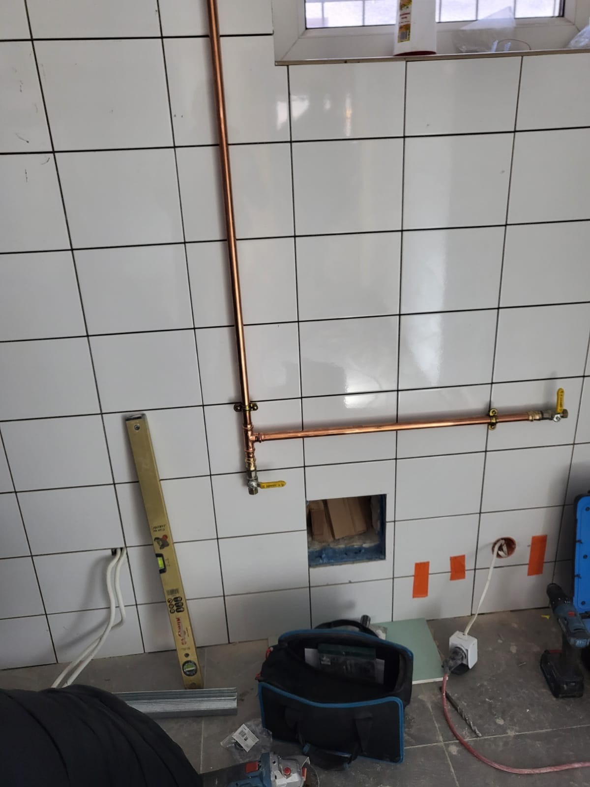 Miedziane rury instalacji wodnej zamontowane na białych kafelkach, widoczne narzędzia, poziomica i torba monterska na podłodze, w tle okno.