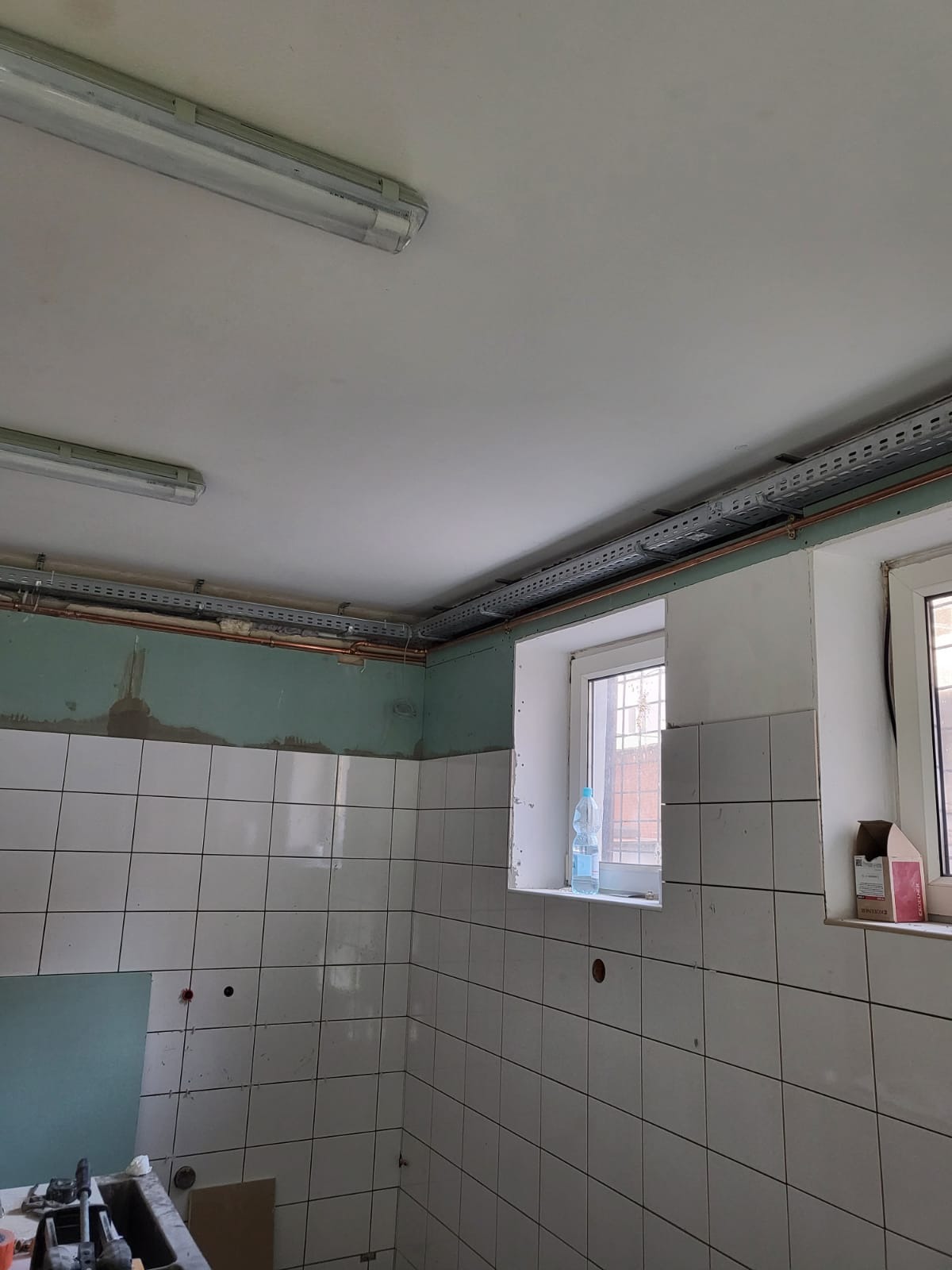 Instalacja rur miedzianych pod sufitem w pomieszczeniu z białymi płytkami, widoczne korytka kablowe, butelka wody na parapecie i fragment narzędzi na dole kadru.