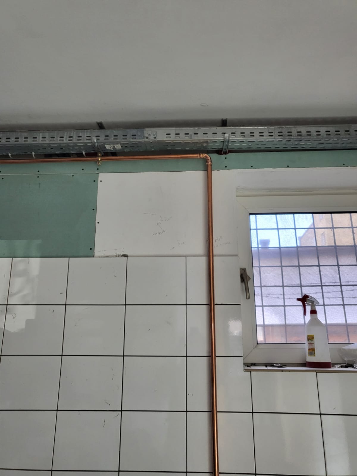 Miedziana rura pionowa z kolankami, zamontowana na tle białych płytek, częściowo zakrytych płytą gipsową i instalacją pod sufitem, widok na okno z kratką i butelką ze spryskiwaczem na parapecie.