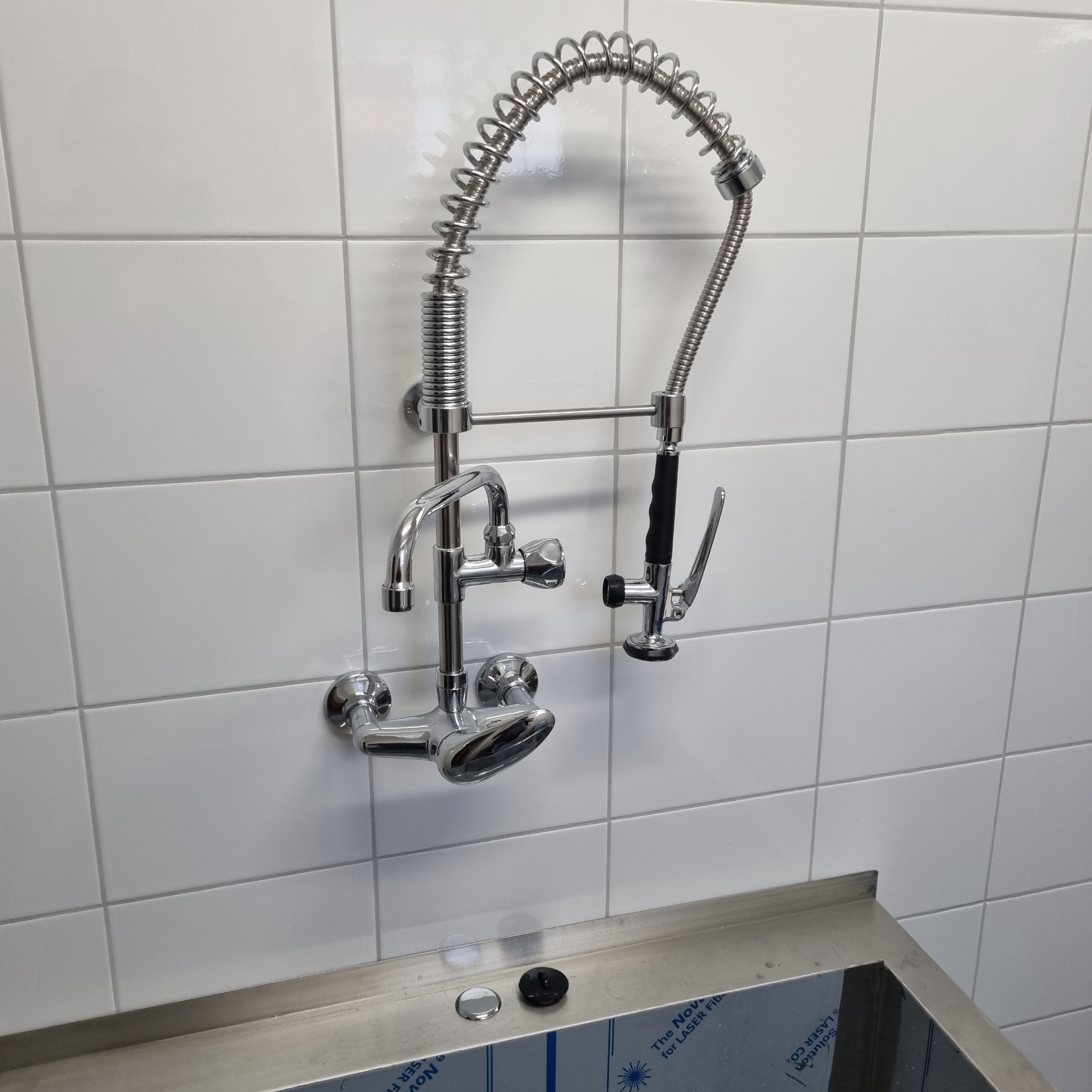 Bateria kuchenna przemysłowa ze sprężynową wylewką i natryskiem, zamontowana na białych płytkach nad stalowym zlewem.