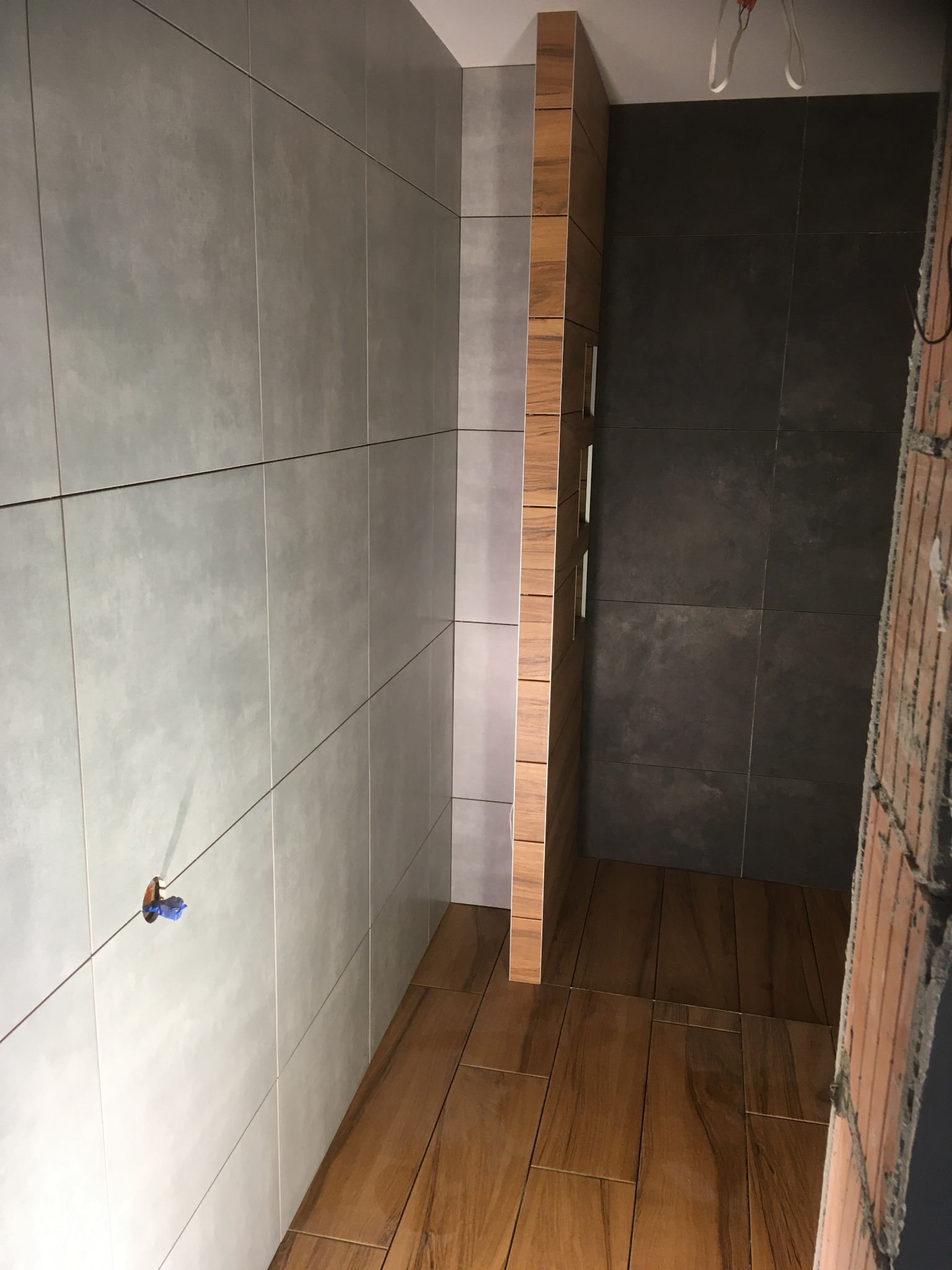 Montaż płytek w łazience: szare płytki ścienne, drewnopodobna podłoga i ciemniejsze płytki w kabinie prysznicowej, widoczne elementy instalacji.