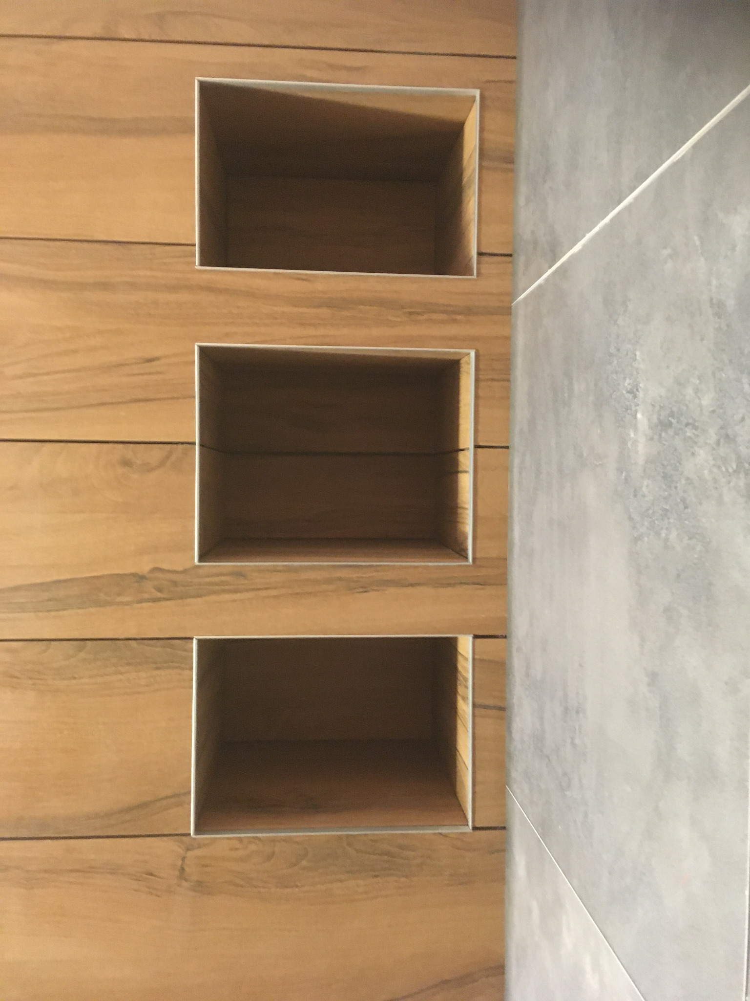 Pionowy układ trzech wnęk z metalowym obramowaniem w ścianie wyłożonej panelami imitującymi drewno, sąsiadującej z szarą, matową płytką ceramiczną.