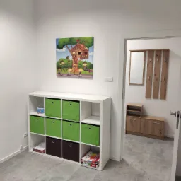 Jasne wnętrze gabinetu z białymi ścianami, regałem z zielonymi i brązowymi pojemnikami, obrazkiem dziecięcego domku na drzewie i widokiem na przedpokój z szafką na buty i wieszakami.